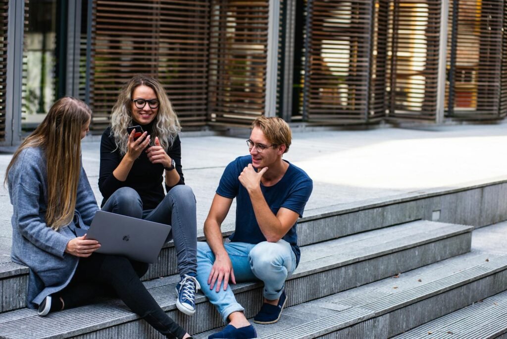 habilidades blandas esenciales, Three young professionals having a friendly chat while sitting on outdoor steps.