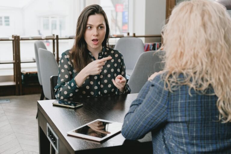 inteligencia emocional en el trabajo, Two women in a lively discussion at a café, with expressive hand gestures and mobile devices on the table.