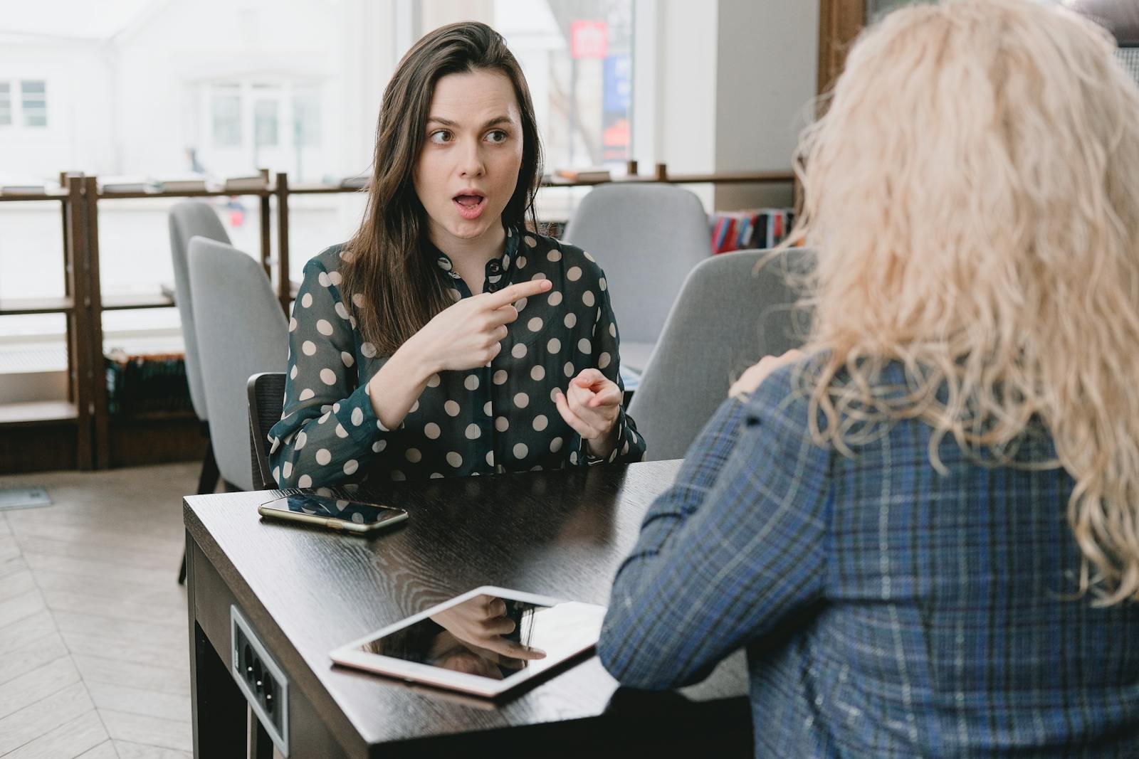 inteligencia emocional en el trabajo, Two women in a lively discussion at a café, with expressive hand gestures and mobile devices on the table.